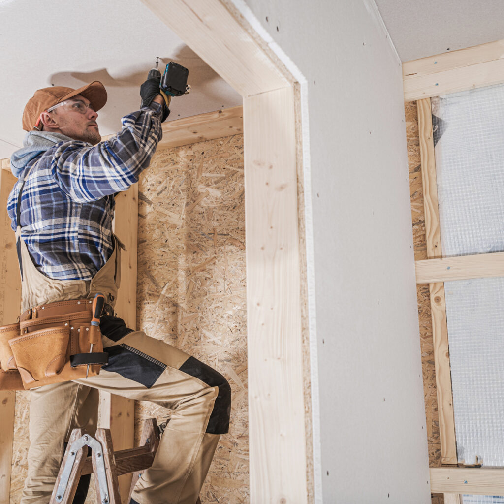 A construction worker in a plaid shirt and cap is installing drywall using a cordless drill atop a ladder.