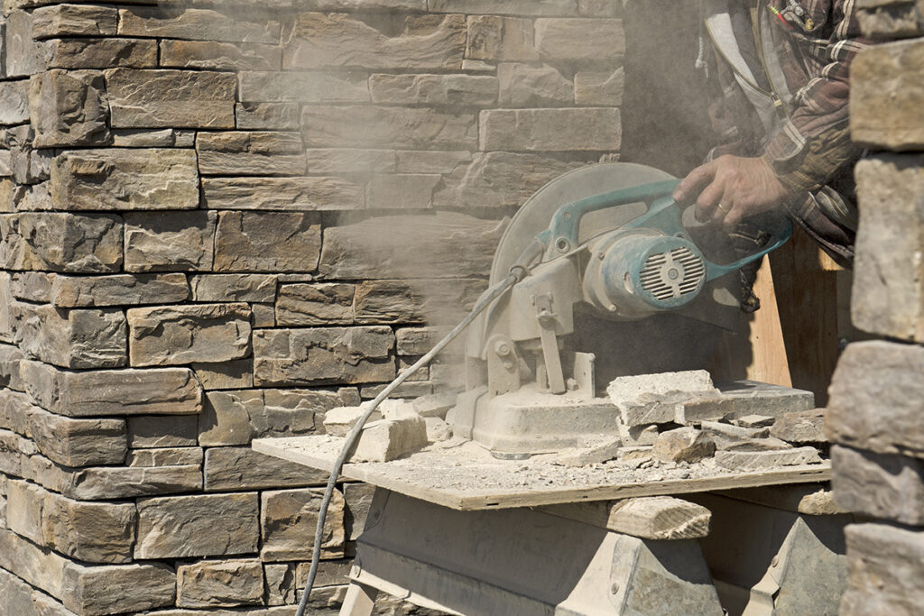 Masonry contractor trimming rock siding with a dry circular saw for home installation.