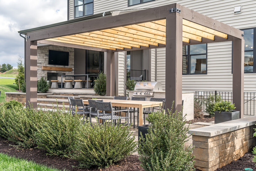 Modern outdoor patio with pergola shade, awning, dining set, grill, and surrounded by greenery.